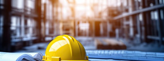 Yellow safety helmet on top of architectural blueprints with a construction site in the background